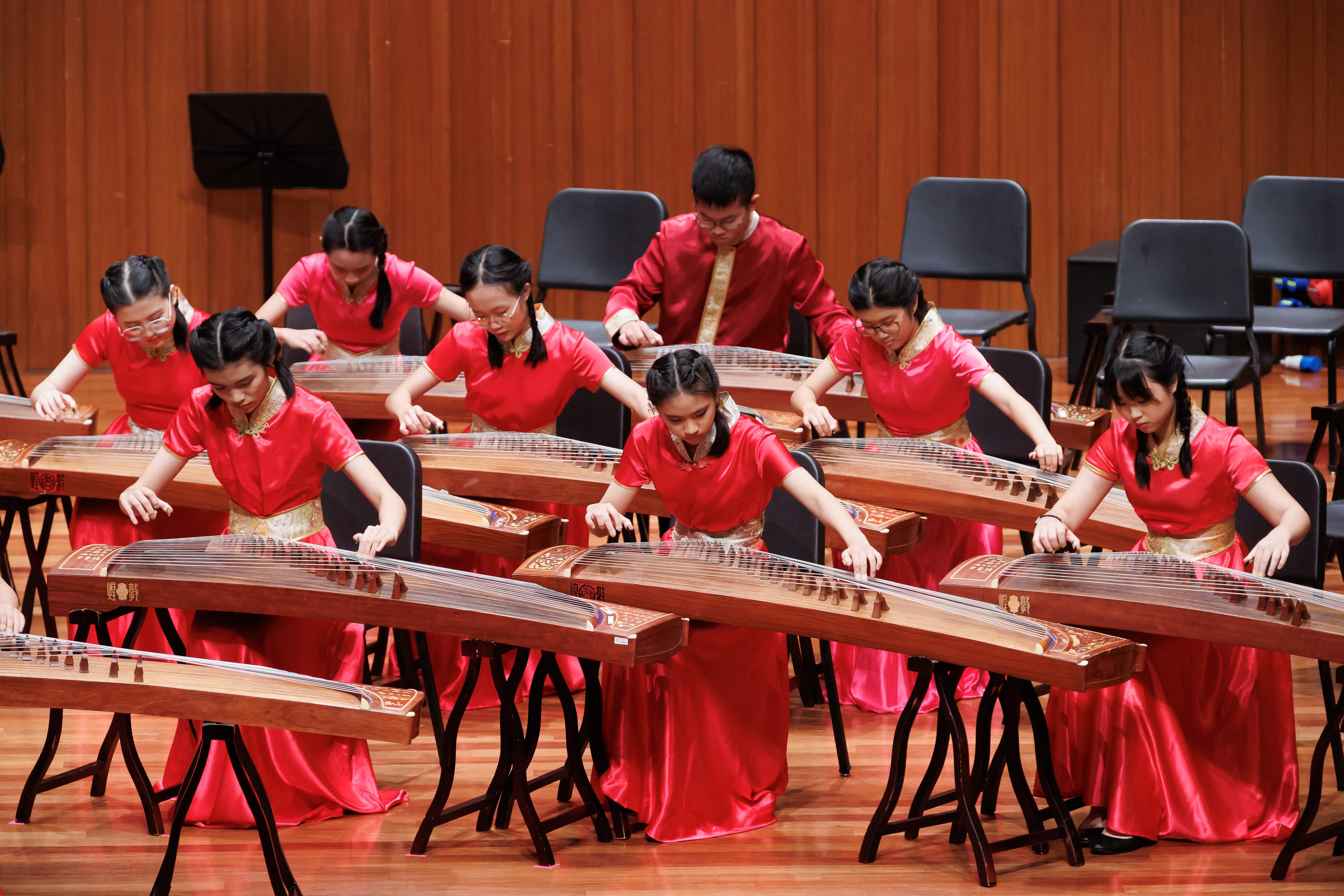 Several young performers in red traditional outfits play guzheng instruments on a wooden stage.